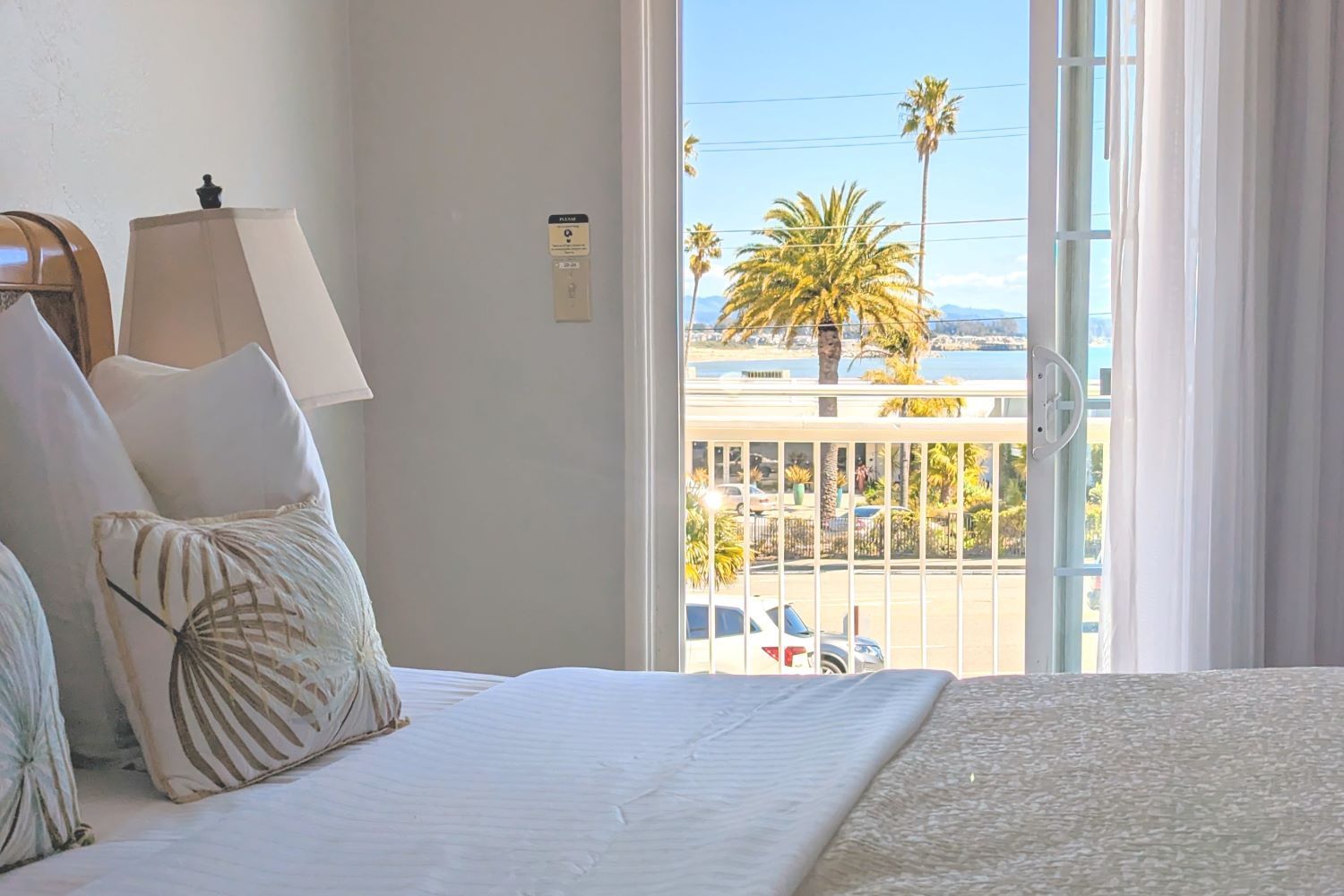 An open sliding glass door with sunshine and palm trees visible outside viewed from behind a neatly made bed