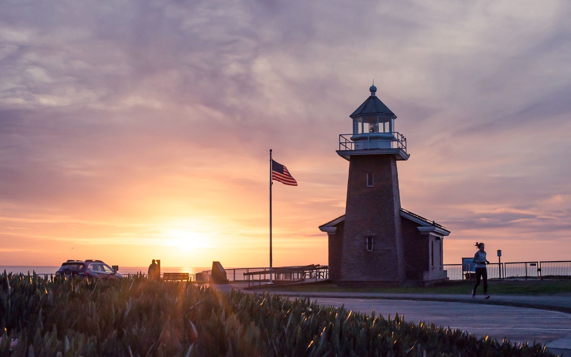Sunset behind Surfing Museum Lighthouse with an American flag flying next to it and people jogging by