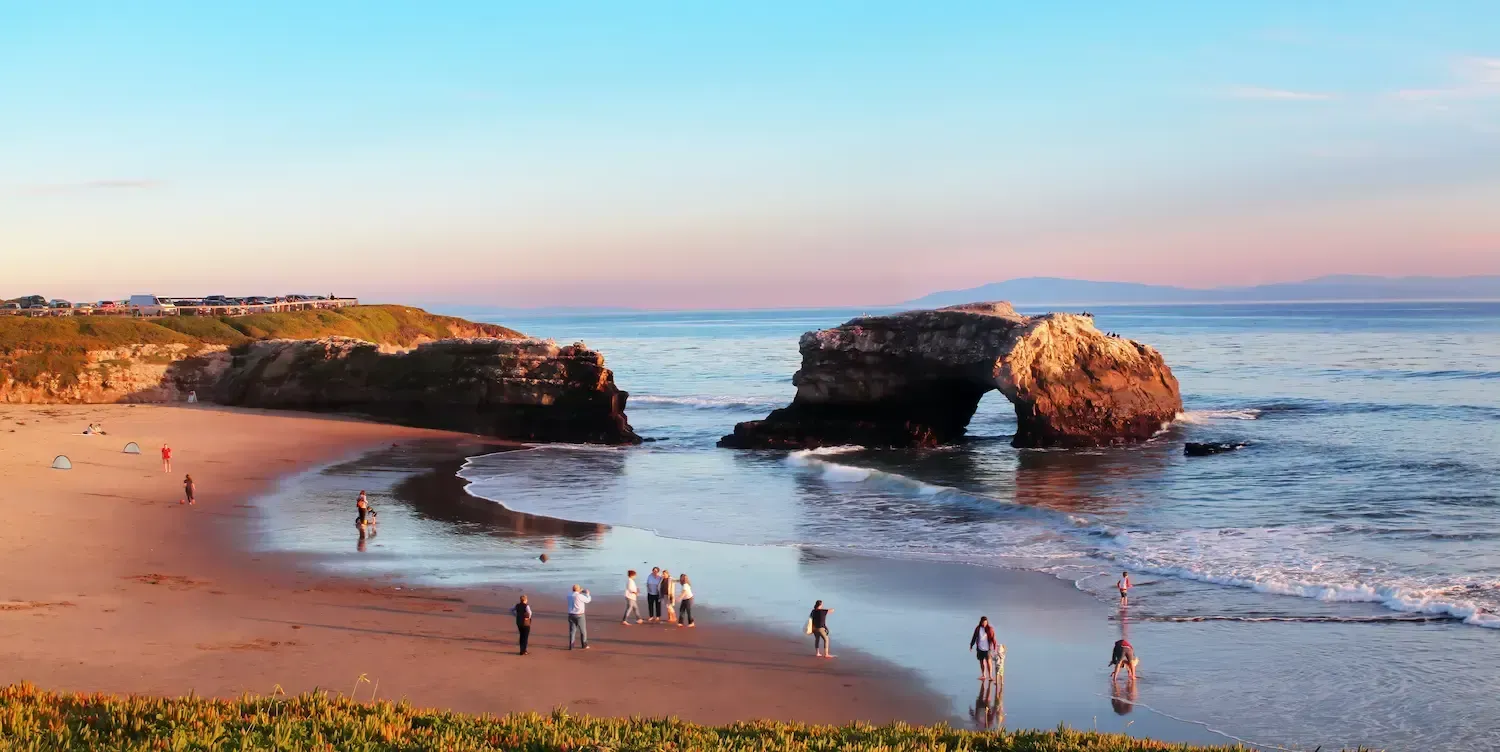 Natural Bridges State Beach with people along the shore in front of the large bridge-like rock formations