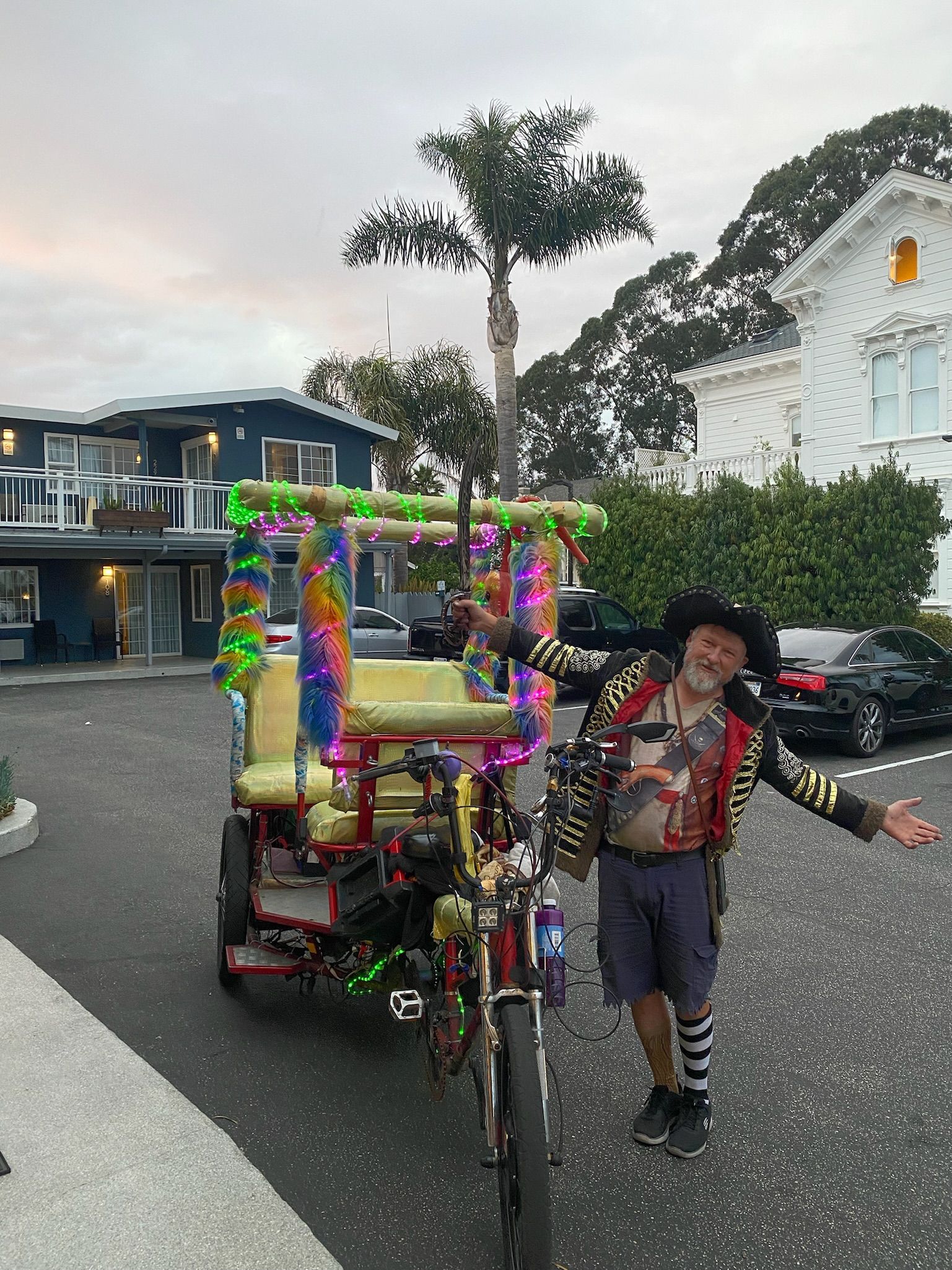 Yellow bicycle with cab in back for riders with a man next to it holding his arms apart in a parking lot with buildings and a palm tree in the background