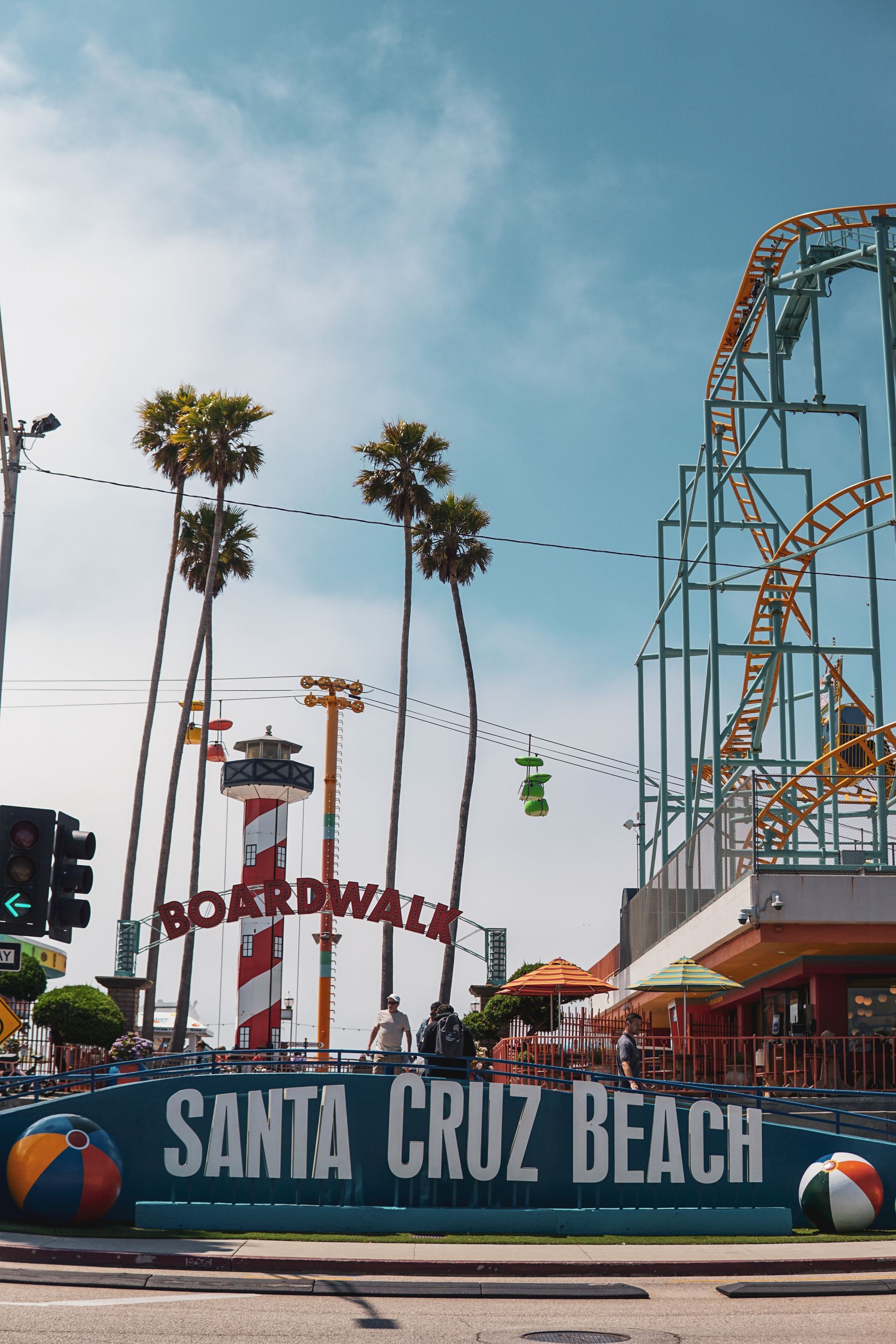 Main entrance to the Santa Cruz Beach Boardwalk with palm trees and a red and white lighthouse in the middle and part of a roller coaster off to the right