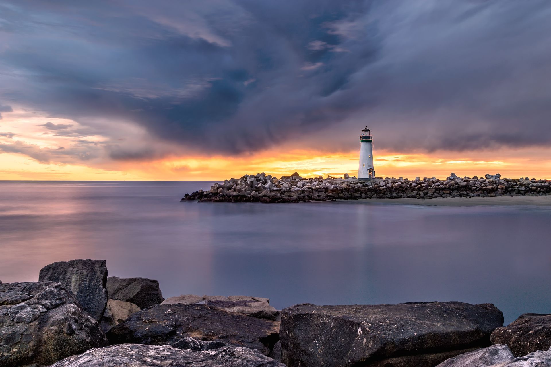 A body of water with large rocks on both sides of it and a lighthouse off in the distance with overcast sunset in the background