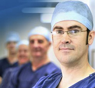 A surgeon wearing glasses and a hat looks at the camera