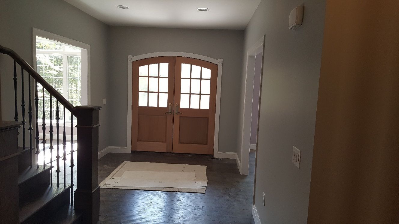 A foyer with a dark wood staircase on the left, light grey walls, and double wooden arched front doors with glass panes.