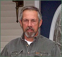 A person with graying hair and a beard, wearing a gray button-down shirt, standing outdoors in front of a house door.