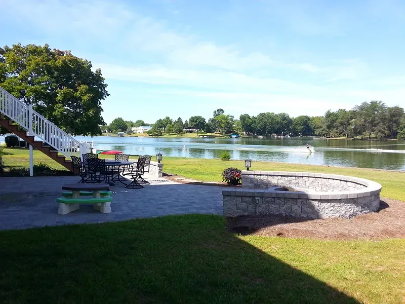 A stone patio with a fire pit, outdoor dining furniture, and stairs overlooking a lake under a clear blue sky.