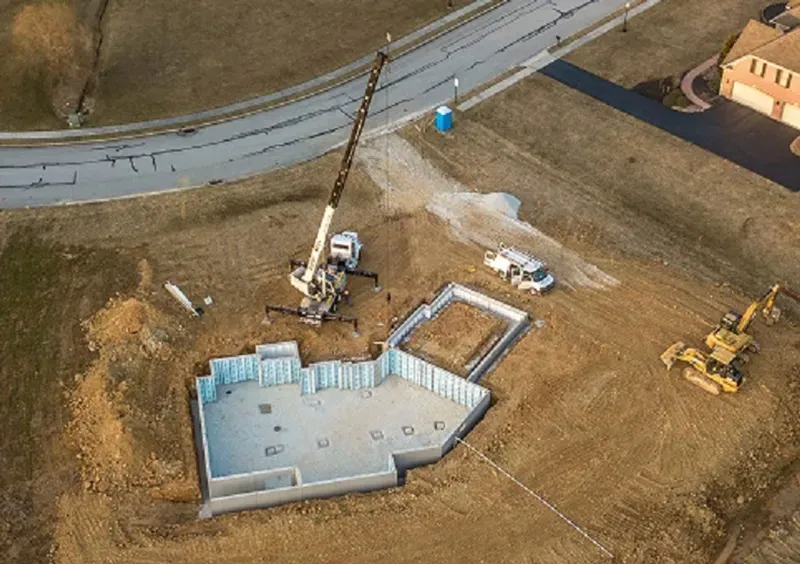 An aerial view of a construction site showing a concrete foundation with blue insulation panels and a mobile crane nearby.