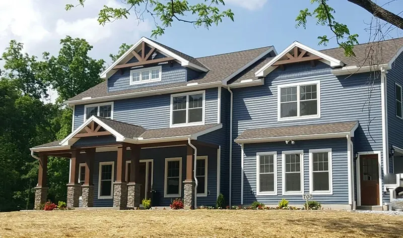 A blue two-story house with a covered porch, stone accents, and a brown shingled roof under a clear blue sky.