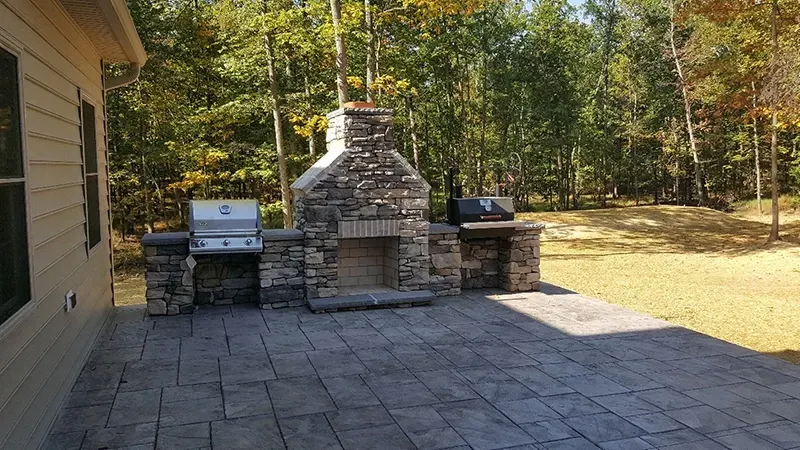 A stone outdoor kitchen with a fireplace and grill on a concrete patio in a backyard with a wooded background.