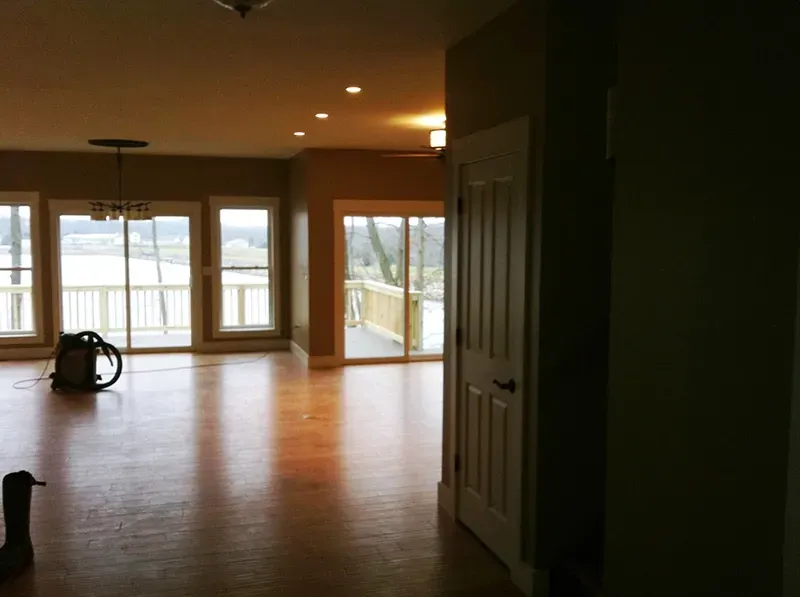 A bright living space with wooden floors, neutral walls, large windows, glass doors leading to a deck, and a vacuum.