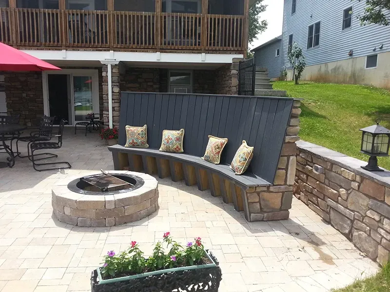 A curved stone-based bench with a dark backrest and floral pillows facing a circular stone fire pit on a patio.