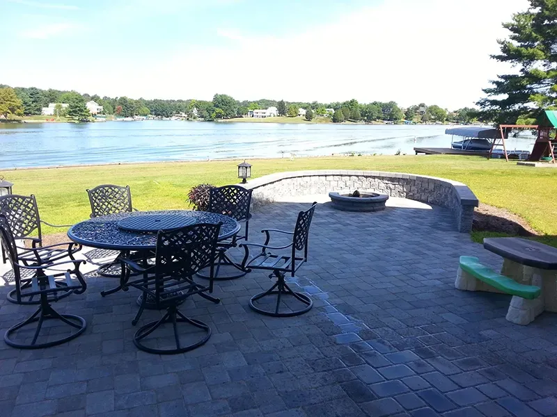 A lakeside patio with a round dining set, stone fire pit, curved seating wall, and a picnic bench overlooking the water.