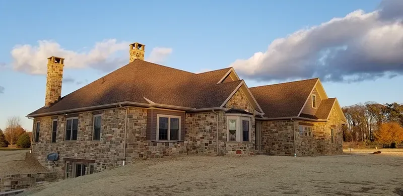 A large, multi-gabled stone house with two chimneys stands on a grassy lot under a partially cloudy blue sky at sunset.