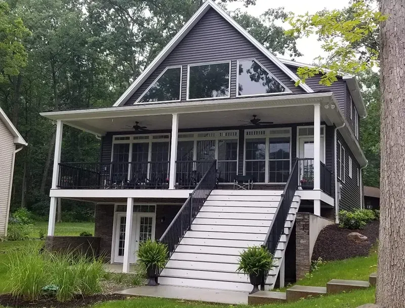A two-story dark gray house with a prominent white staircase, upper deck, and large windows set in a wooded area.