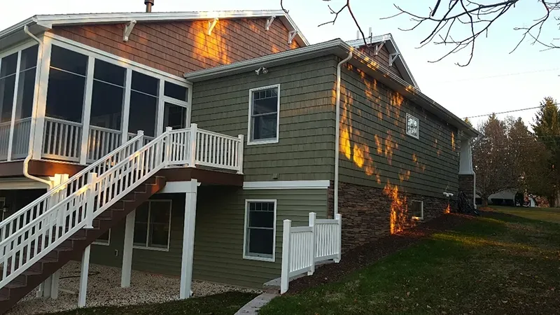 A side view of a two-story home with olive green siding, a raised deck with white railings, and a stone foundation.