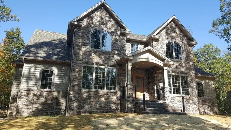 Two-story stone house with a gabled roof, dark window frames, and a covered front entryway surrounded by trees.