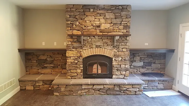 A large stone fireplace with a central hearth and wood mantels on either side of a room with neutral-colored walls.