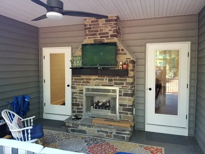 A covered outdoor patio with a stone fireplace, mounted television, two white glass doors, and a decorative area rug.