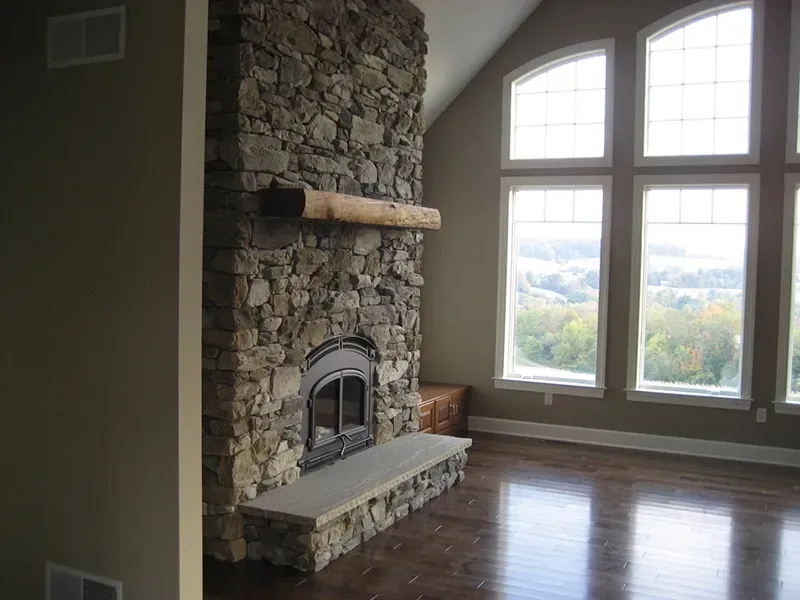 A tall, natural stone fireplace with a thick wooden mantel stands in a room with hardwood floors and arched windows.