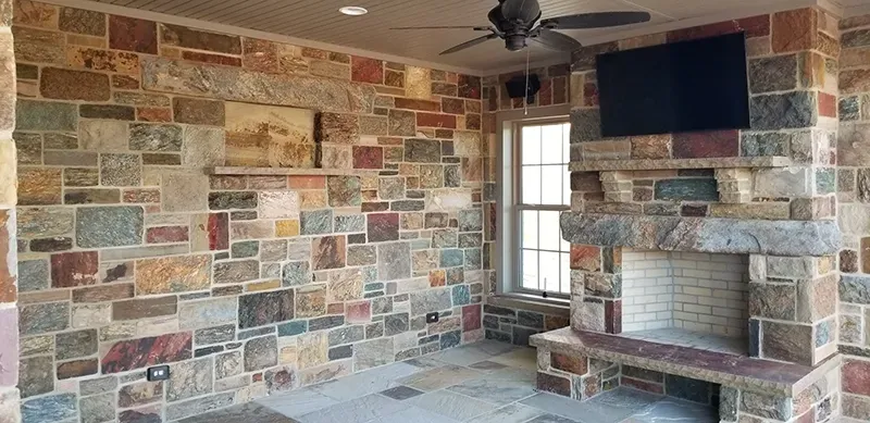 An outdoor living space with a stone wall, a fireplace with a mounted television above it, and a ceiling fan.