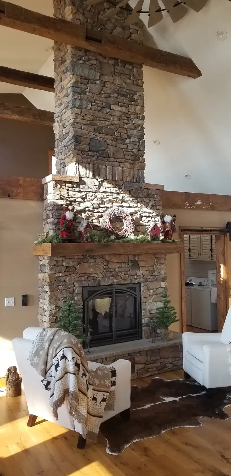 A tall stone fireplace in a rustic room with exposed wooden beams, two white chairs, and a brown cowhide rug.