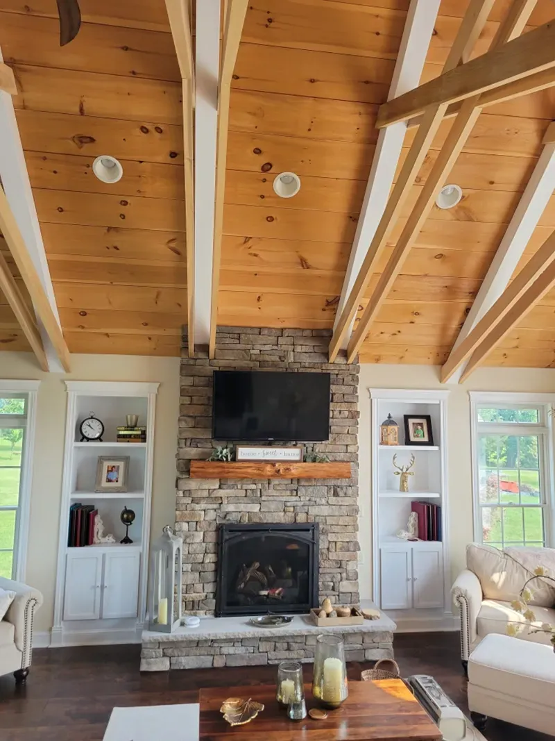 A living room features a stone fireplace with a television above it, flanked by two white bookshelves under a wood ceiling.
