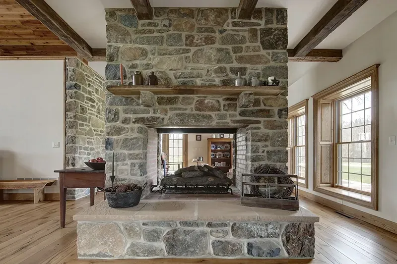 A double-sided stone fireplace with a wooden mantel, set in a room with hardwood floors and exposed wooden ceiling beams.