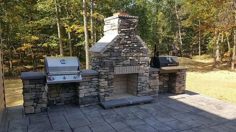 An outdoor stone fireplace flanked by a built-in stainless steel grill and a smaller cooking unit on a stone patio.