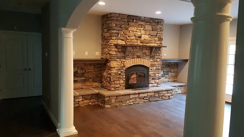 A floor-to-ceiling stone fireplace with a wooden mantel, flanked by built-in stone shelves, seen through white columns.