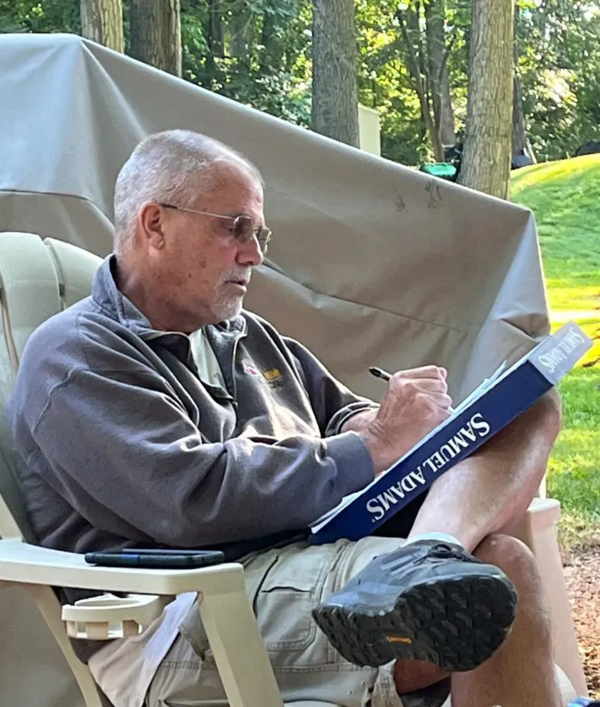 A person sits in a chair outdoors, writing on a Samuel Adams branded pad while wearing glasses and a gray pullover.