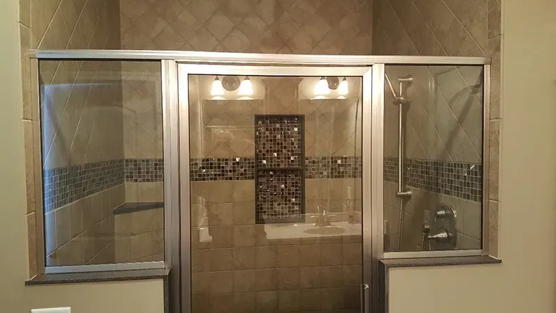 A framed glass shower door with side panels, featuring tan square tiles and a dark decorative accent border.