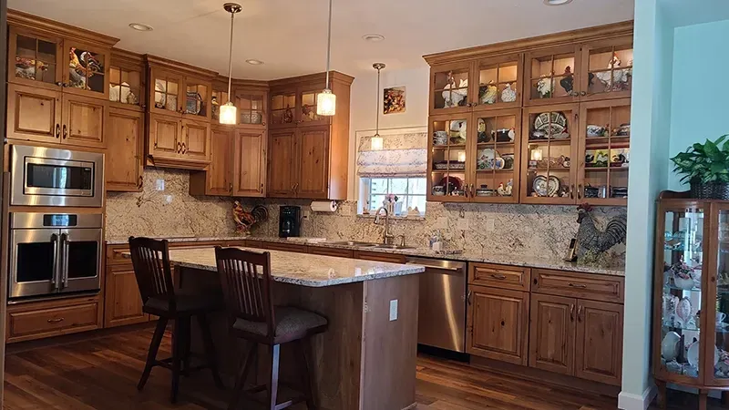 A kitchen with warm wood cabinets, granite countertops, a center island with two stools, and glass-front upper cabinets.