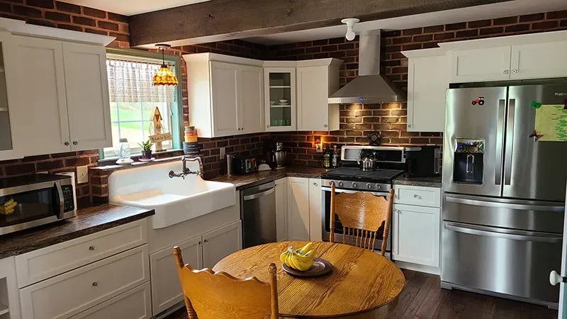 Kitchen with brick walls, white cabinets, stainless steel appliances, farmhouse sink, and a wooden table in the center.