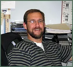 A man with a beard and glasses wearing a grey and black striped polo shirt, smiling while sitting at a desk.