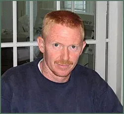A man with short reddish-brown hair and a mustache wearing a dark blue sweatshirt in front of a window.