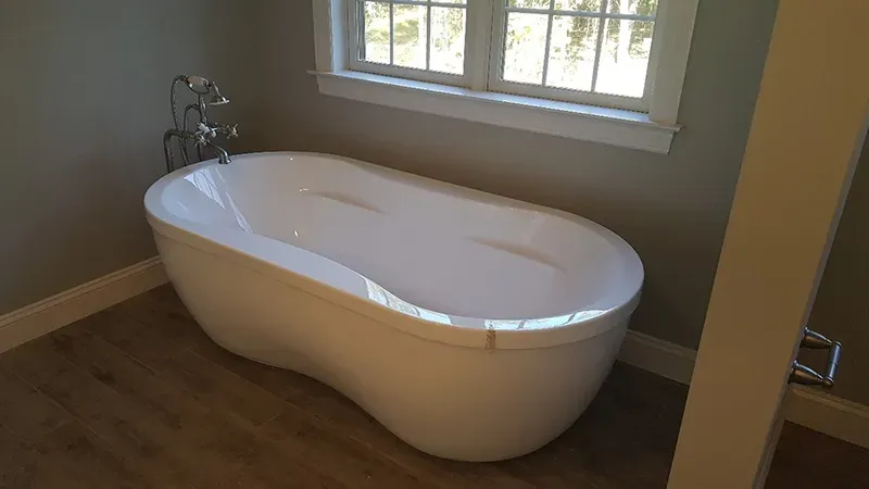 A white freestanding bathtub sits on a wood-look floor in front of a window in a room with grey walls.