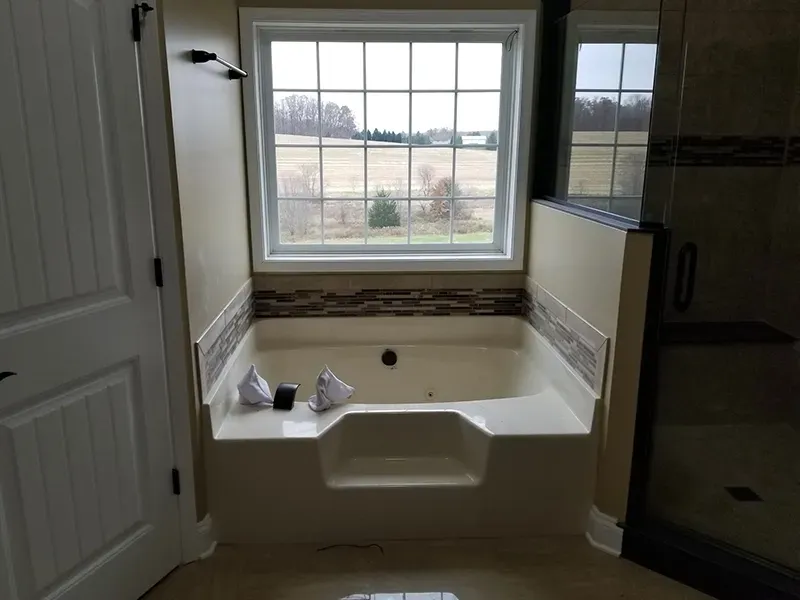 A beige built-in bathtub centered under a window in a bathroom with tiled walls and a door on the left.