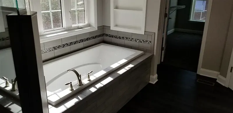 A built-in white bathtub with a chrome faucet in a bathroom featuring gray tiles and dark wood flooring.