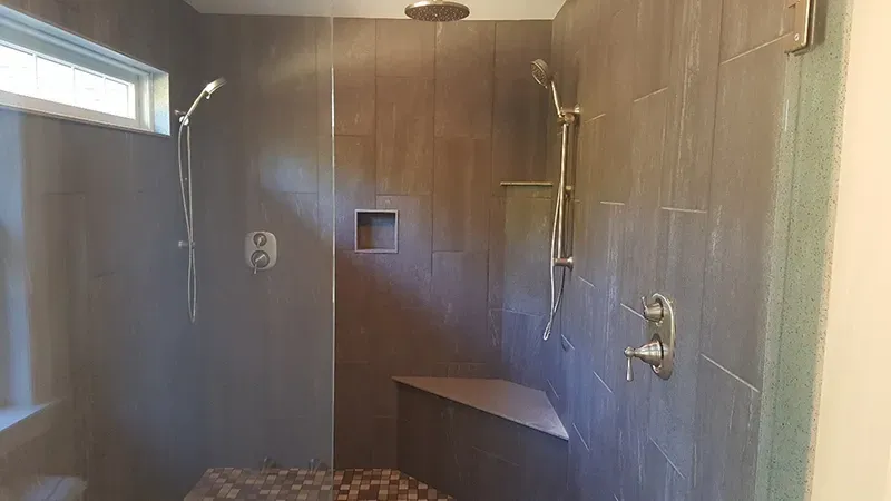 A modern walk-in shower with dark grey tiles, a corner bench, a rain shower head, and two handheld shower wands.