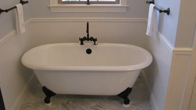A white clawfoot bathtub with black hardware stands against a wall with white beadboard wainscoting and blue paint.