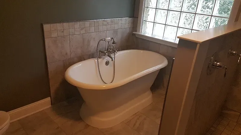 A white freestanding bathtub with a silver faucet stands in a bathroom corner with tiled walls and a glass block window.