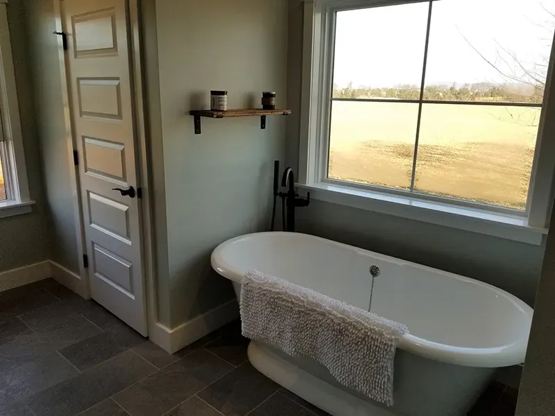 A freestanding white bathtub sits by a large window in a room with light green walls and dark gray tile flooring.