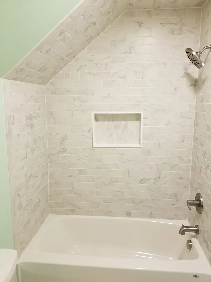 Bathtub and shower with light gray marble-look wall tiles, a built-in recessed niche, and modern brushed nickel fixtures.