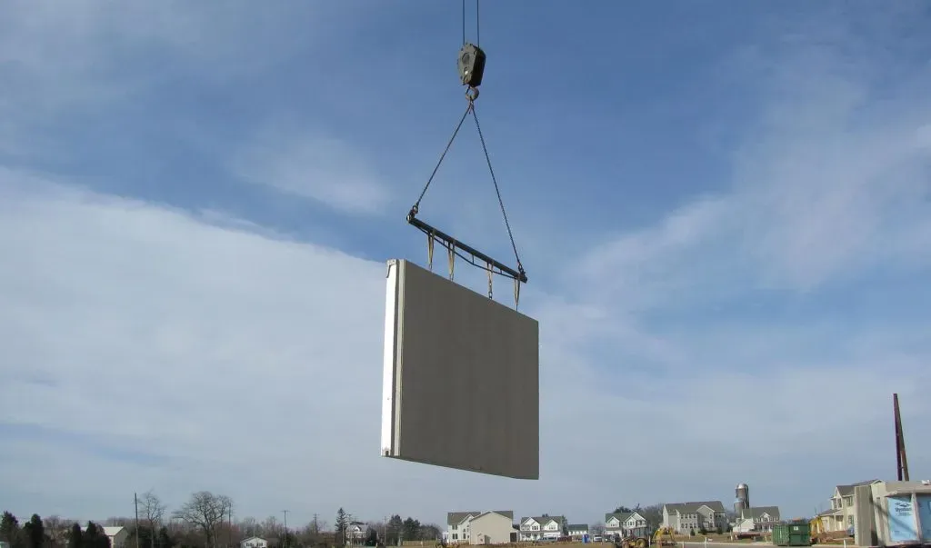 A large rectangular concrete panel hangs suspended in the air from a construction crane against a blue sky.
