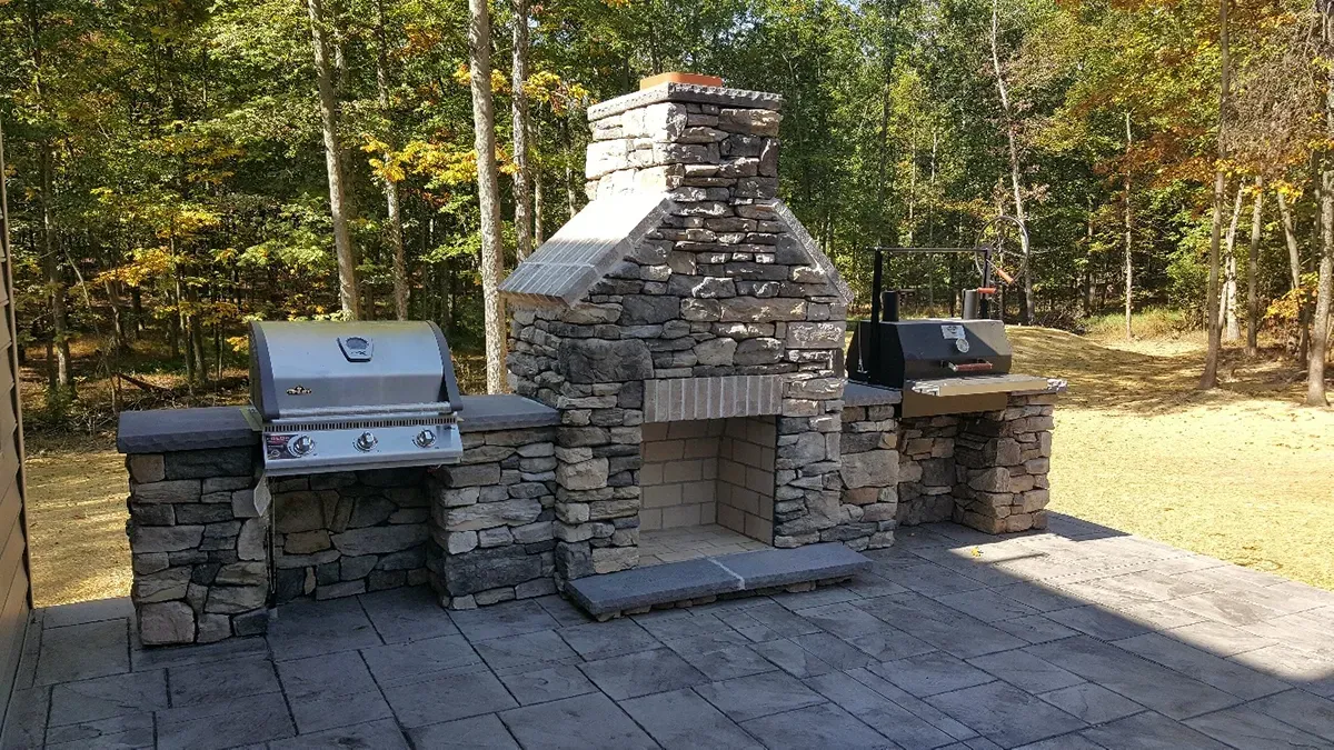 A stone outdoor kitchen featuring a central fireplace flanked by a stainless steel grill and a countertop in a wooded area.