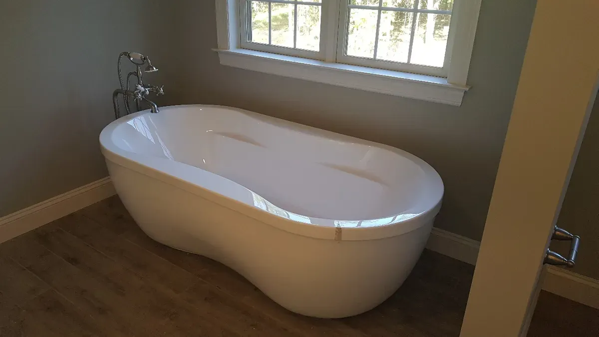 A white, oval-shaped freestanding bathtub sits on a wooden floor in front of a white window in a grey room.