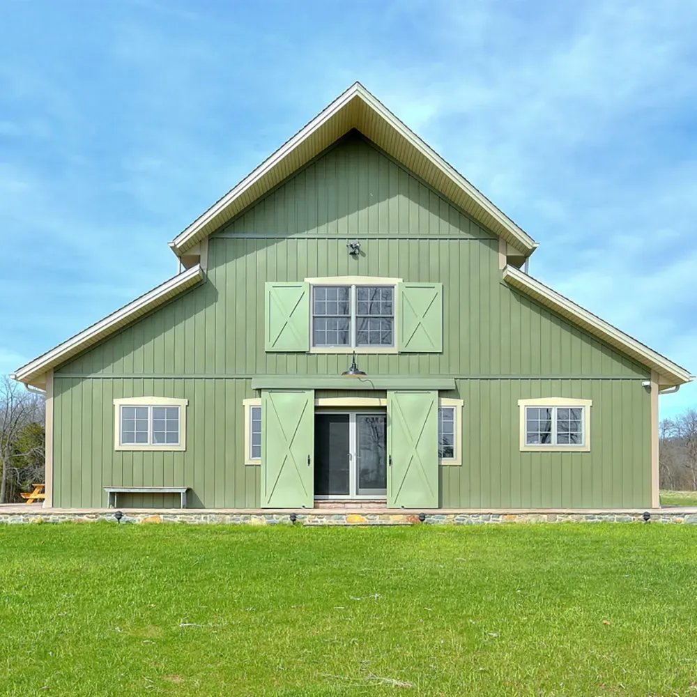 A green barn with large sliding doors, a centered window, and a gabled roof set against a blue sky above a grassy lawn.