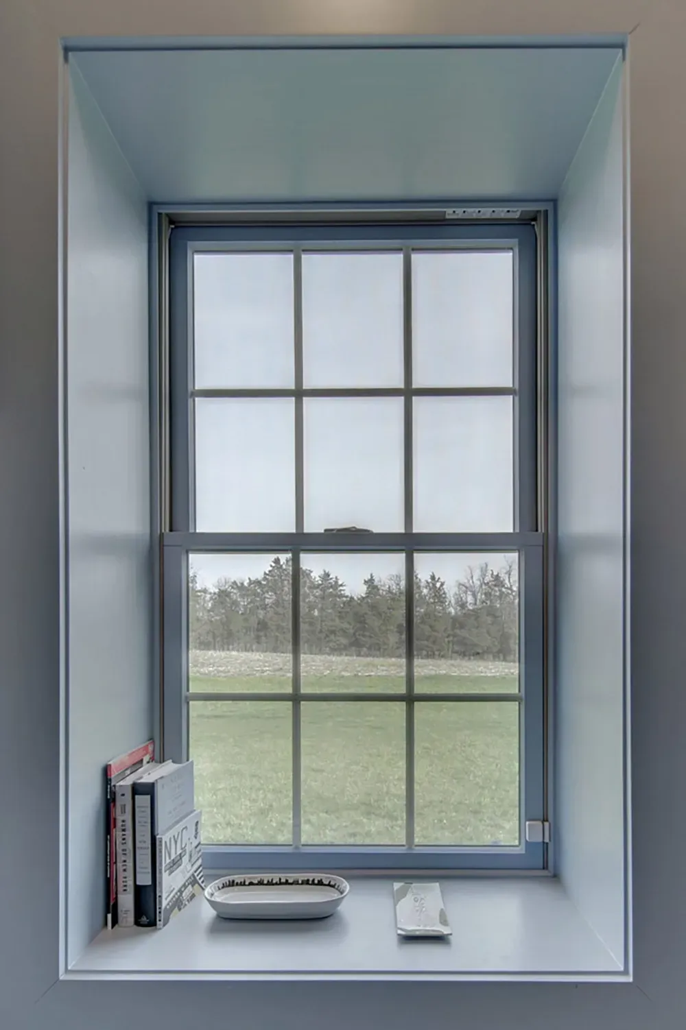 A light blue recessed window frame overlooks a grassy field, with books and small trays sitting on the white sill.
