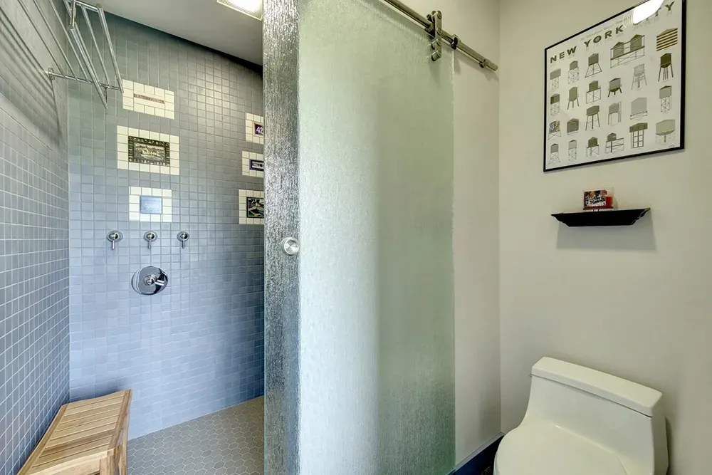 A modern bathroom with a tiled walk-in shower behind a frosted sliding door next to a white toilet and wall art.