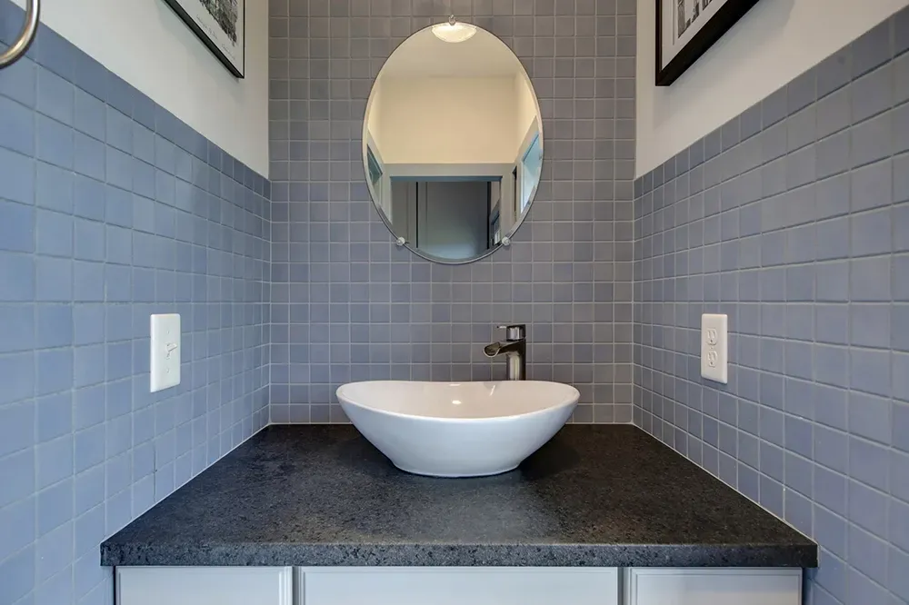 A white oval vessel sink sits on a dark stone countertop against a wall of square, slate-blue tiles with a round mirror.